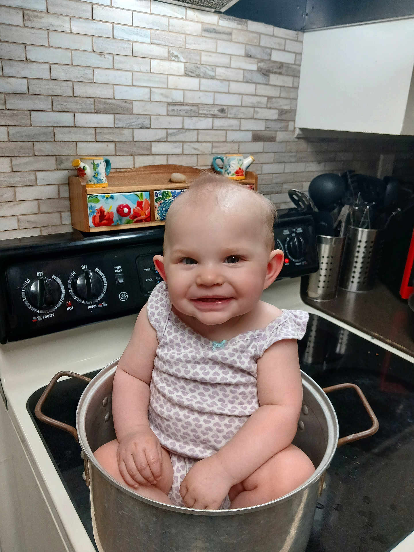 Hartley is registered to the contest to win money with this photo: baby, child, kitchen, pot, stove, smiling, cooking_utensils, countertop, tile_wall, appliance, cute, infant, sitting, metal_pot, domestic, indoors, playful, happy, person, wearing_clothes
