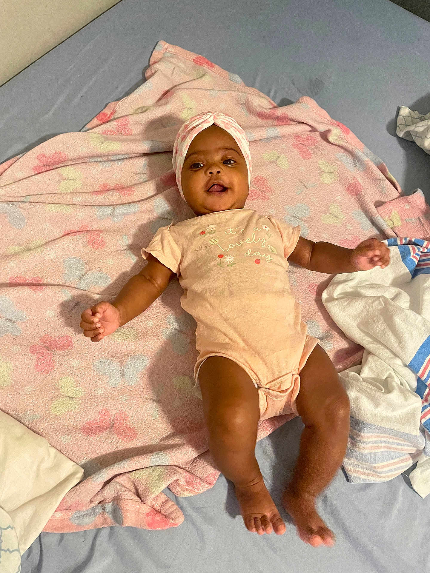 Kemani is registered to the contest to win money with this photo: baby, infant, blanket, head_wrap, onesie, bed, smiling, lying_down, cute, child, soft_texture, patterned_blanket, indoors, happy, person, skin, feet, hands, face, comfort