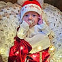 baby, infant, christmas, holiday, hat, knitted_blanket, fairy_lights, pacifier, red_clothing, cozy, festive, decorations, floor, wooden_floor, tree_branch, ornament, cute, portrait, indoors, celebration