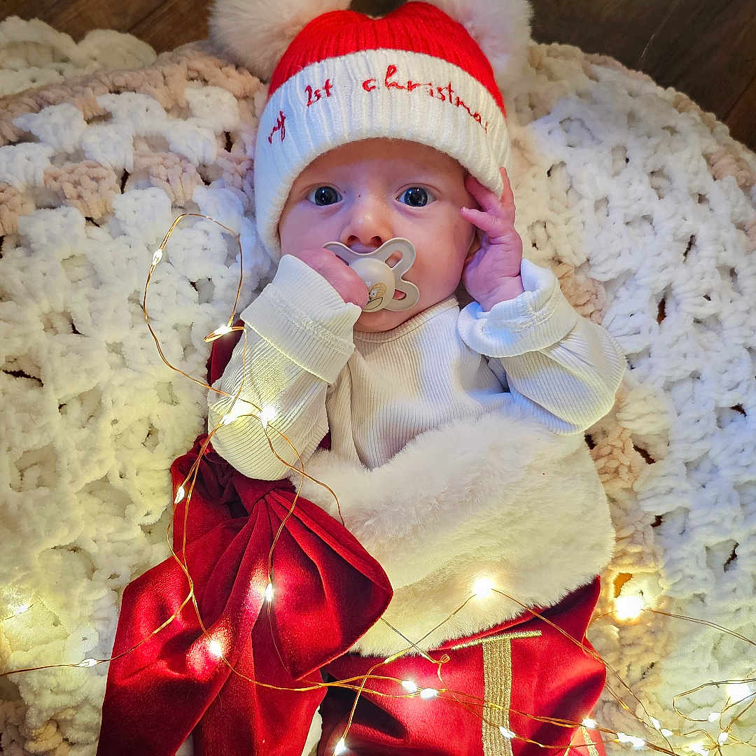 Lochie is registered to the contest to win money with this photo: baby, celebration, christmas, cozy, cute, decorations, fairy_lights, festive, floor, hat, holiday, indoors, infant, knitted_blanket, ornament, pacifier, portrait, red_clothing, tree_branch, wooden_floor