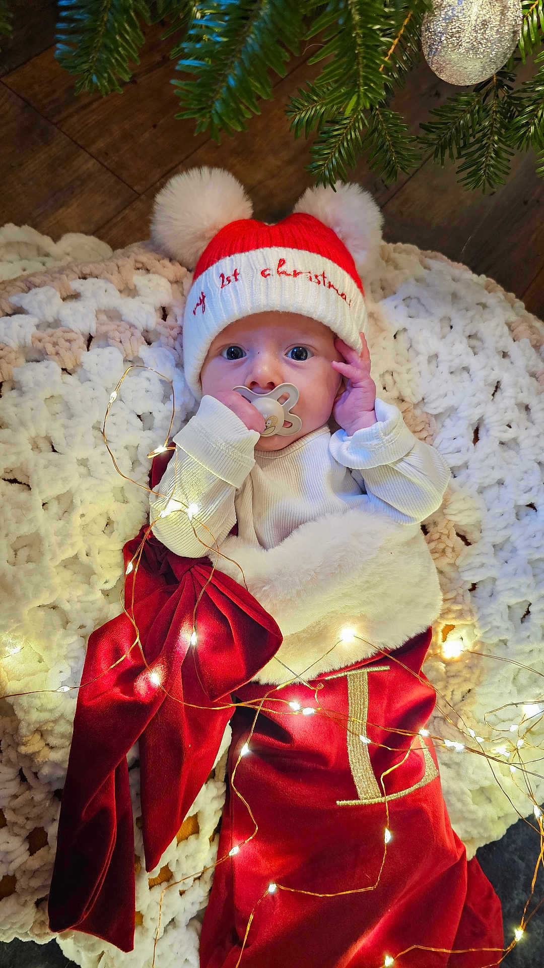 Lochie is registered to the contest to win money with this photo: baby, infant, christmas, holiday, hat, knitted_blanket, fairy_lights, pacifier, red_clothing, cozy, festive, decorations, floor, wooden_floor, tree_branch, ornament, cute, portrait, indoors, celebration