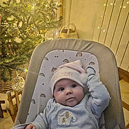 Emy participe au concours pour gagner de l'argent avec cette photo : baby, basket, blue_clothing, chair, child, christmas_tree, cozy, cute, elephant_design, festive, hat, holiday, indoors, infant, lights, ornaments, seatbelt, smiling, warm_lighting, wood_floor