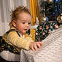 celebration, child, christmas_ornaments, christmas_tree, curious, curly_hair, decorations, dress, face, festive, holiday, home, indoor, person, reaching_hand, soft_lighting, tablecloth, toddler, wrapped_presents, yellow_sweater