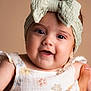 baby, child, headband, bow, smiling, face, hand, floral_dress, infant, portrait, cute, happy, closeup, skin, person, soft_background, apparel, young_child, studio_photo, expression