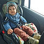 blue_sweater, car_interior, car_seat, child, child_safety, cozy, curious_expression, floral_cushion, indoor, knitwear, portrait, safety_harness, seatbelt, shoes, sitting, striped_hat, toddler, vehicle, window, young_child