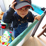 Léo a rejoint le concours — aidez-le/la à gagner de superbes lots ! cap, child, clothing, colorful, cute, denim_overalls, floor, furniture, hand, hat, holding, indoor, person, play_area, playpen, portrait, smiling, toddler, toys, young_child