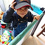 cap, child, clothing, colorful, cute, denim_overalls, floor, furniture, hand, hat, holding, indoor, person, play_area, playpen, portrait, smiling, toddler, toys, young_child