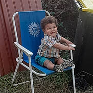 Léo a rejoint le concours — aidez-le/la à gagner de superbes lots ! casual_clothing, chair, child, curly_hair, daylight, grass, happy, nature, outdoor, person, plaid_shirt, red_wall, shoes, shorts, sitting, smiling, sun_emblem, toddler, trash_bin, yellow_lid