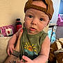 arm, baby, blanket, blue_eyes, cap, casual_clothing, child, curious, face, green_tank_top, hat, holding, indoor, jeep_graphic, person, portrait, soft_furniture, tattoo, tiger, toy