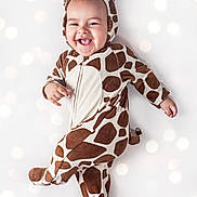 Aaron is registered to the contest to win money with this photo: baby, child, giraffe_costume, onesie, smiling, tongue_out, happy, cute, infant, playful, costume, animal_print, portrait, face, feet, hand, light_background, bokeh, soft_lighting, indoor
