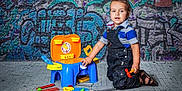 Aaron joined the competition — help win amazing prizes! toddler, child, toy_tools, playset, denim_overalls, striped_shirt, kneeling, graffiti_wall, urban_background, colorful, playtime, floor, plastic_toys, curious, young_child, indoors, casual_clothing, bright_colors, portrait, happy