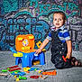 toddler, child, toy_tools, playset, denim_overalls, striped_shirt, kneeling, graffiti_wall, urban_background, colorful, playtime, floor, plastic_toys, curious, young_child, indoors, casual_clothing, bright_colors, portrait, happy