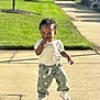 toddler, child, smiling, sidewalk, sunlight, green_lawn, pants, sneakers, short_sleeve_shirt, outdoor, daytime, curly_hair, walking, cute, playful, suburban, shadow, happy, person, casual_clothing