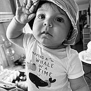 Abran is registered to the contest to win money with this photo: baby, child, hat, bucket_hat, shirt, text_on_clothing, indoor, portrait, black_and_white, cute, wide_eyes, hand_raised, expression, face, person, closeup, infant, home, casual_clothing, smile
