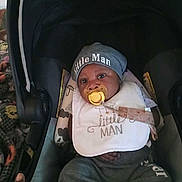 Jaiquan is registered to the contest to win money with this photo: baby, infant, child, car_seat, pacifier, hat, bib, clothing, portrait, sitting, indoors, vehicle, dark_background, face, eyes, newborn, casual, infantwear, text_on_clothing, photography