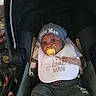 baby, infant, child, car_seat, pacifier, hat, bib, clothing, portrait, sitting, indoors, vehicle, dark_background, face, eyes, newborn, casual, infantwear, text_on_clothing, photography