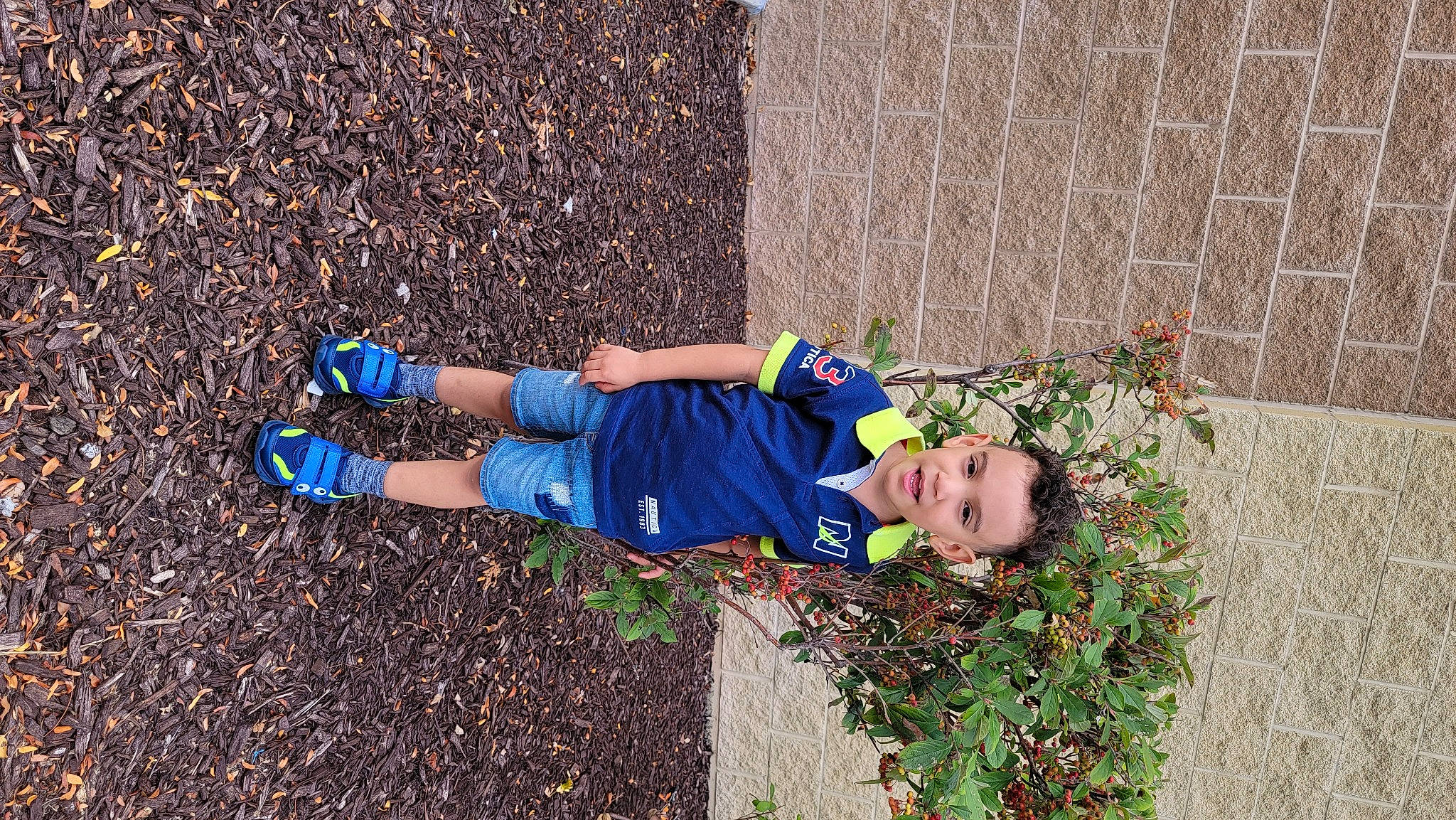 Dylan is registered to the contest to win money with this photo: asphalt, brick, electric_blue, grass, grass_family, groundcover, hat, human_leg, leisure, people_in_nature, person, plant, road_surface, sidewalk, sitting, sneakers, soil, street_fashion, t_shirt, toddler