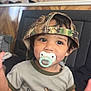 toddler, child, pacifier, hat, camo_hat, shirt, duck, moose, sitting, indoor, couch, cute, baby, young_child, clothing, face, person, smiling, headwear, casual