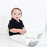 toddler, child, black_polo, white_pants, gray_socks, disco_ball, sitting, indoor, white_background, curious, wide_eyes, minimalist, cute, portrait, young_child, holding_object, studio_lighting, casual_clothing, innocent, baby