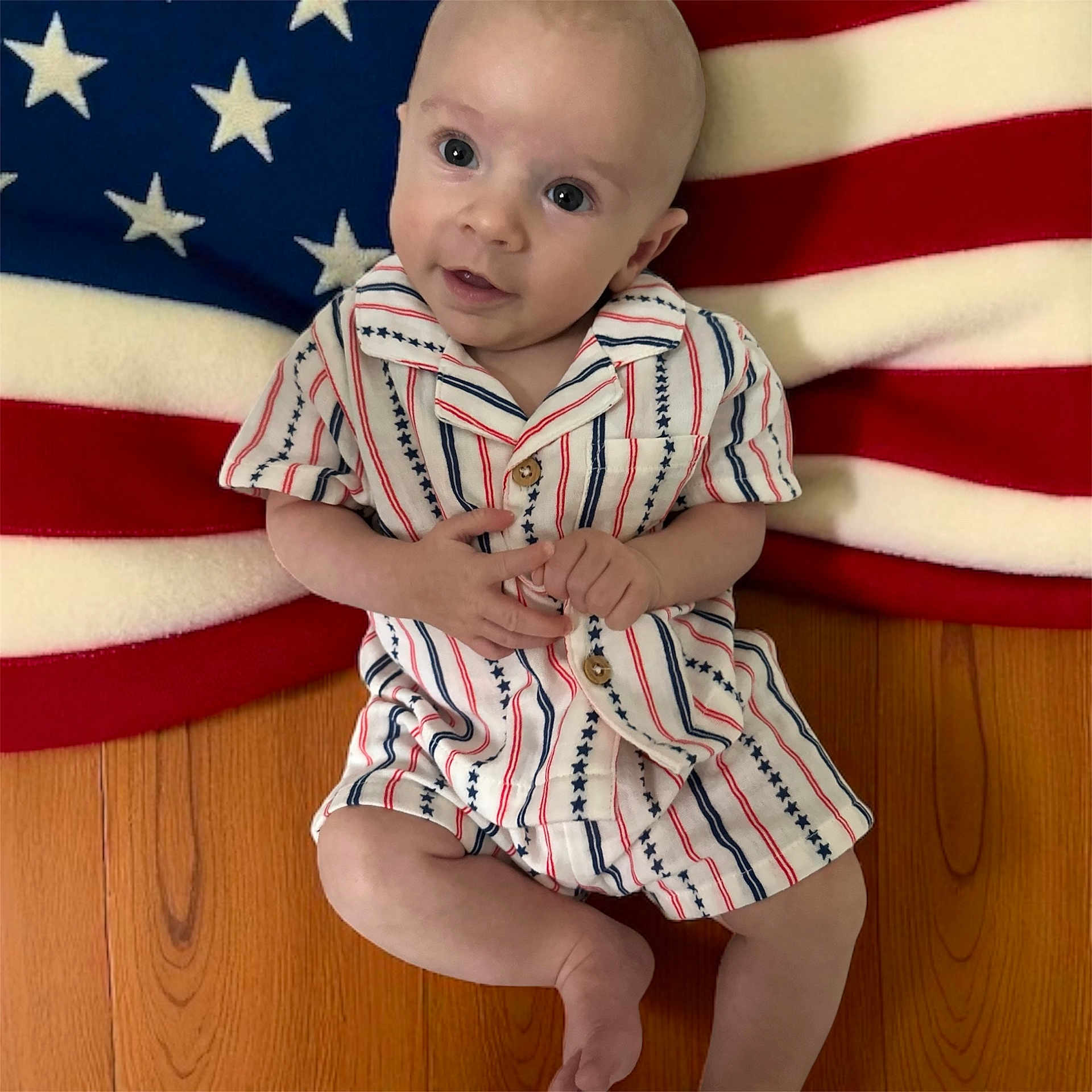 Randy is registered to the contest to win money with this photo: baby, infant, child, floor, blanket, american_flag, romper, clothing, wood, pattern, stars, stripes, cute, portrait, indoor, looking, hands, sitting, face, expression