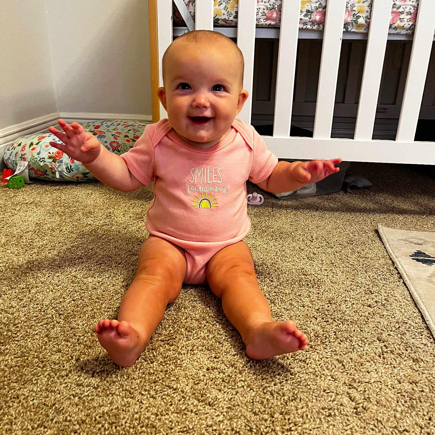 Emelia joined the competition — help win amazing prizes! arms_outstretched, baby, carpet, child, crib, cushion, cute, face, floor, floral_bedding, happy, head, indoor, infant, nursery, person, pink_onesie, room, smiling, toes