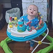 Christopher is registered to the contest to win money with this photo: baby, child, walker, toy, blue_onesie, floor, wooden_floor, cabinet, trash_can, indoor, foot, face, person, furniture, colorful, plastic, playtime, curious, standing, home