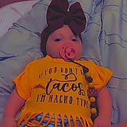 Keely is registered to the contest to win money with this photo: baby, pacifier, headband, bow, blanket, yellow_shirt, taco, shorts, clothing, infant, lying_down, indoors, cute, fringe, colorful, funny_text, child, person, comfort, relaxing