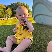 Vivian is registered to the contest to win money with this photo: baby, child, keys, yellow_clothing, sunflower_pattern, outdoor, playground, slide, grass, green_turf, blue_sky, sunlight, sitting, barefoot, cute, infant, playing, chewing, happy, nature