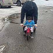 Merlin is registered to the contest to win money with this photo: adult, black_clothing, blue_jacket, car, child, cold_weather, footwear, hoodie, outdoor, parking_lot, pavement, person, residential_area, snow, toy_vehicle, tricycle, truck, wet_ground, winter, young_child