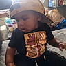 toddler, child, cap, backwards_cap, black_tshirt, peace_sign, sitting, bed, indoor, furniture, bottle, container, curtain, shelf, plastic_bag, clothing, person, young_child, casual, room