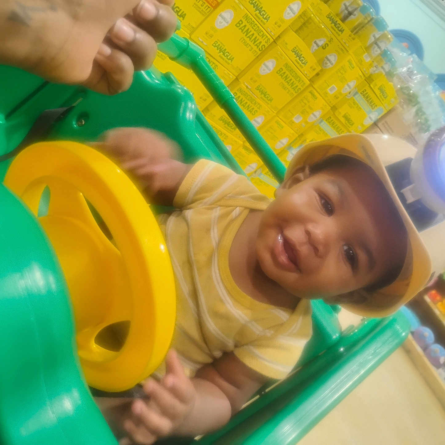 Bobby is registered to the contest to win money with this photo: baby, child, smiling, toy_car, yellow_shirt, helmet, light, indoor, store, boxes, toys, colorful, hand, face, person, happy, playful, green_toy, yellow_steering_wheel, infant