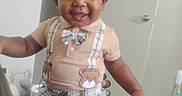 Bobby is registered to the contest to win money with this photo: toddler, child, smiling, indoor, plaid_shorts, suspenders, teddy_bear, bow_tie, happy, person, face, curly_hair, short_sleeve_shirt, door, hand, floor, wall, baby, clothing, holding
