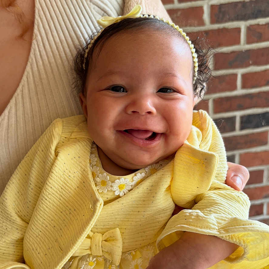 Joyful joined the competition — help win amazing prizes! adult_hand, baby, brick_wall, child, clothing, cute, daisy_pattern, fabric, face, hand, happy, headband, holding, indoor, infant, person, portrait, smiling, texture, yellow_dress
