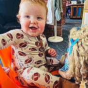 Liam joined the competition — help win amazing prizes! baby, infant, smiling, face, blue_eyes, orange_chair, pajamas, football_pattern, doll, toy, brick_wall, indoor, carpet, happy, curly_hair, blue_bow, hand, mouth_open, portrait, cute