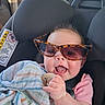 baby, child, car_seat, sunglasses, blanket, pacifier, pink_shirt, smiling, tongue_out, cute, infant, seat_belt, striped_blanket, toddler, indoors, person, happy, car_interior, head, fun