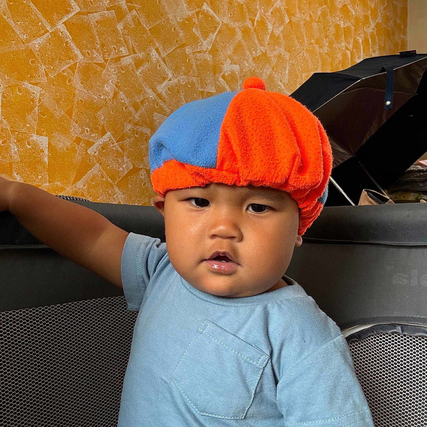 Elijah is registered to the contest to win money with this photo: arm, blue, blue_shirt, casual_clothing, child, expression, face, furniture, hat, home, indoor, mesh, orange, person, playpen, serious, standing, texture, toddler, wall