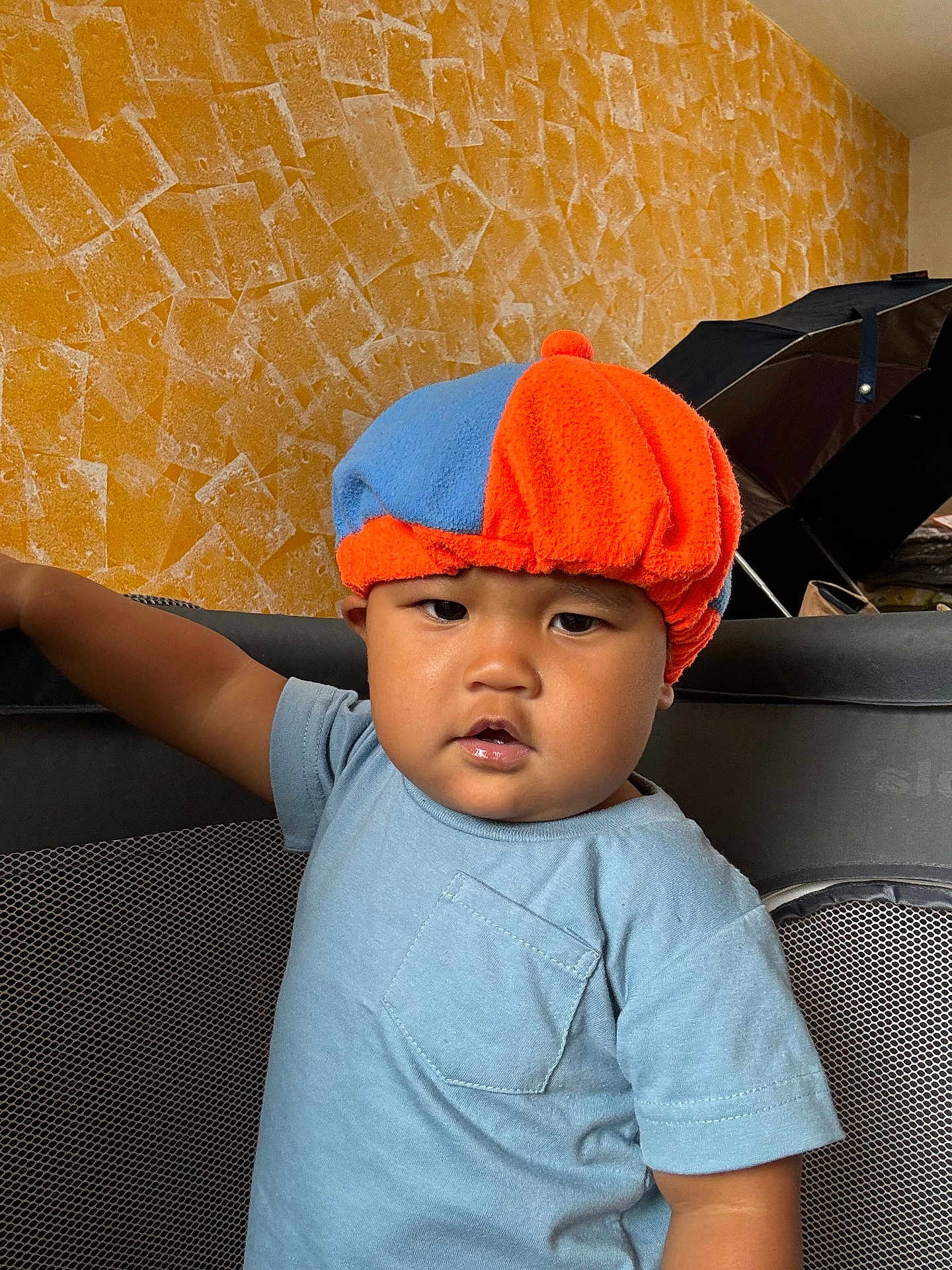 Elijah is registered to the contest to win money with this photo: toddler, child, hat, blue_shirt, playpen, indoor, person, face, expression, serious, orange, blue, wall, texture, arm, standing, home, furniture, mesh, casual_clothing