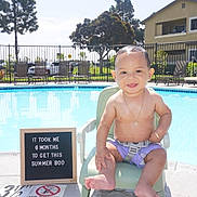 Javier is registered to the contest to win money with this photo: baby, child, pool, swimming_pool, chair, smiling, outdoor, sunny, summer, sign, text, purple_shorts, harness, happy, water, residential_building, tree, fence, footwear_free, person