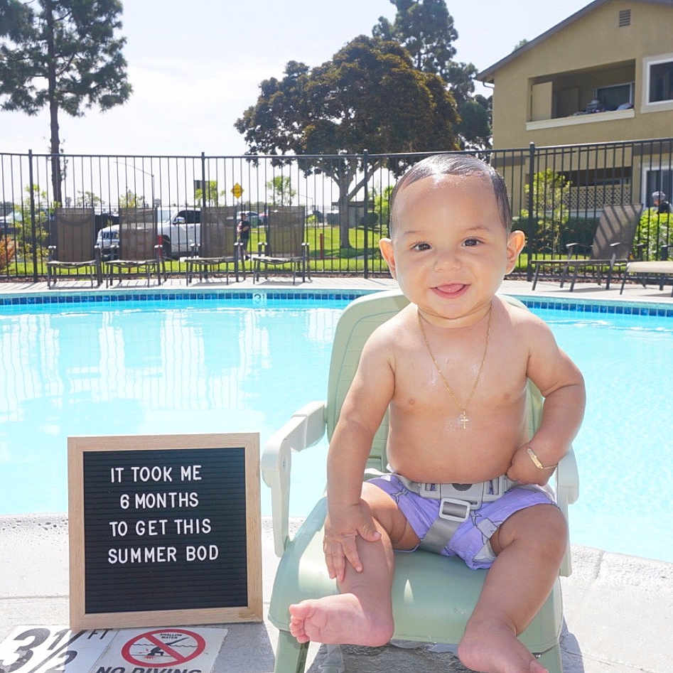 Javier is registered to the contest to win money with this photo: baby, child, pool, swimming_pool, chair, smiling, outdoor, sunny, summer, sign, text, purple_shorts, harness, happy, water, residential_building, tree, fence, footwear_free, person