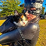 person, cat, costume, calico_cat, outdoor, daylight, greenery, tree, grass, blue_sky, portrait, animal, pet, black_costume, leather, face, holding, closeup, sunlight, nature