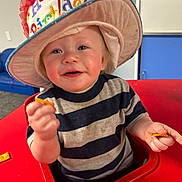 Gunner is registered to the contest to win money with this photo: baby, child, infant, smiling, happy, birthday_hat, party, high_chair, red_chair, striped_shirt, food, crackers, snack, indoors, classroom, portrait, close_up, seated, colorful, playful