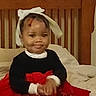 toddler, child, bow, headwear, red_dress, black_top, bed, wooden_headboard, indoors, smile, hands, seated, clothing, portrait, cute, baby, young_child, furniture, pajamas, home