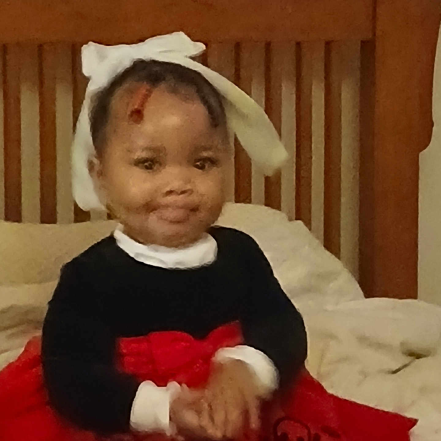 Maya is registered to the contest to win money with this photo: toddler, child, bow, headwear, red_dress, black_top, bed, wooden_headboard, indoors, smile, hands, seated, clothing, portrait, cute, baby, young_child, furniture, pajamas, home