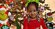 Za'Layah is registered to the contest to win money with this photo: child, christmas_tree, grinch, holiday, festive, lights, ornaments, pajamas, beads, braids, smile, red_clothing, green_bows, indoor, decorations, celebration, seasonal, happy, sitting, toy