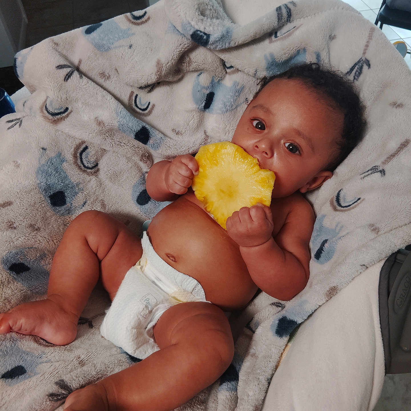 Ethan is registered to the contest to win money with this photo: baby, infant, diaper, pineapple, fruit, blanket, curly_hair, cute, hand, foot, indoor, floor, tile_floor, person, skin, relaxing, lying_down, holding, eating, home