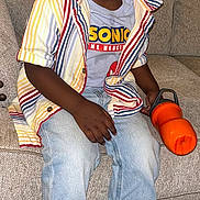 Zaccai is registered to the contest to win money with this photo: toddler, child, smiling, sitting, couch, striped_shirt, jeans, sneakers, water_bottle, indoor, carpet, cute, portrait, casual_clothing, happy, person, short_hair, fashion, colorful, comfortable