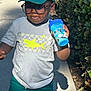 child, boy, glasses, cap, dinosaur, tshirt, shorts, sippy_cup, outdoor, sidewalk, sunlight, shadow, bushes, green, playful, tongue_out, casual_clothing, summer, person, smile