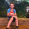 children, baby, boy, siblings, aquarium, fish, underwater, portrait, sitting, holding, smiling, family, wooden_bench, striped_shirt, orange_clothing, sneakers, indoor, zoo, aquatic_life, photography