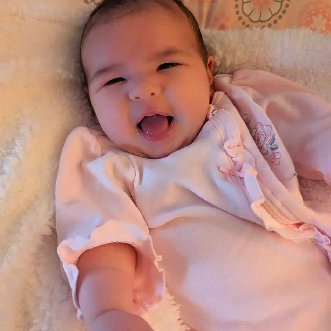 Cheyenne is registered to the contest to win money with this photo: baby, infant, smiling, pink_clothing, blanket, reaching_hand, happy, cute, lying_down, cozy, soft_texture, indoor, portrait, child, young_baby, human, face, warm_colors, patterned_background, sleepwear