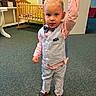 toddler, child, boy, indoor, carpet, clothing, vest, bow_tie, striped_pants, shoes, crib, adult_hand, room, wall, floor, furniture, portrait, standing, cute, holding_hand
